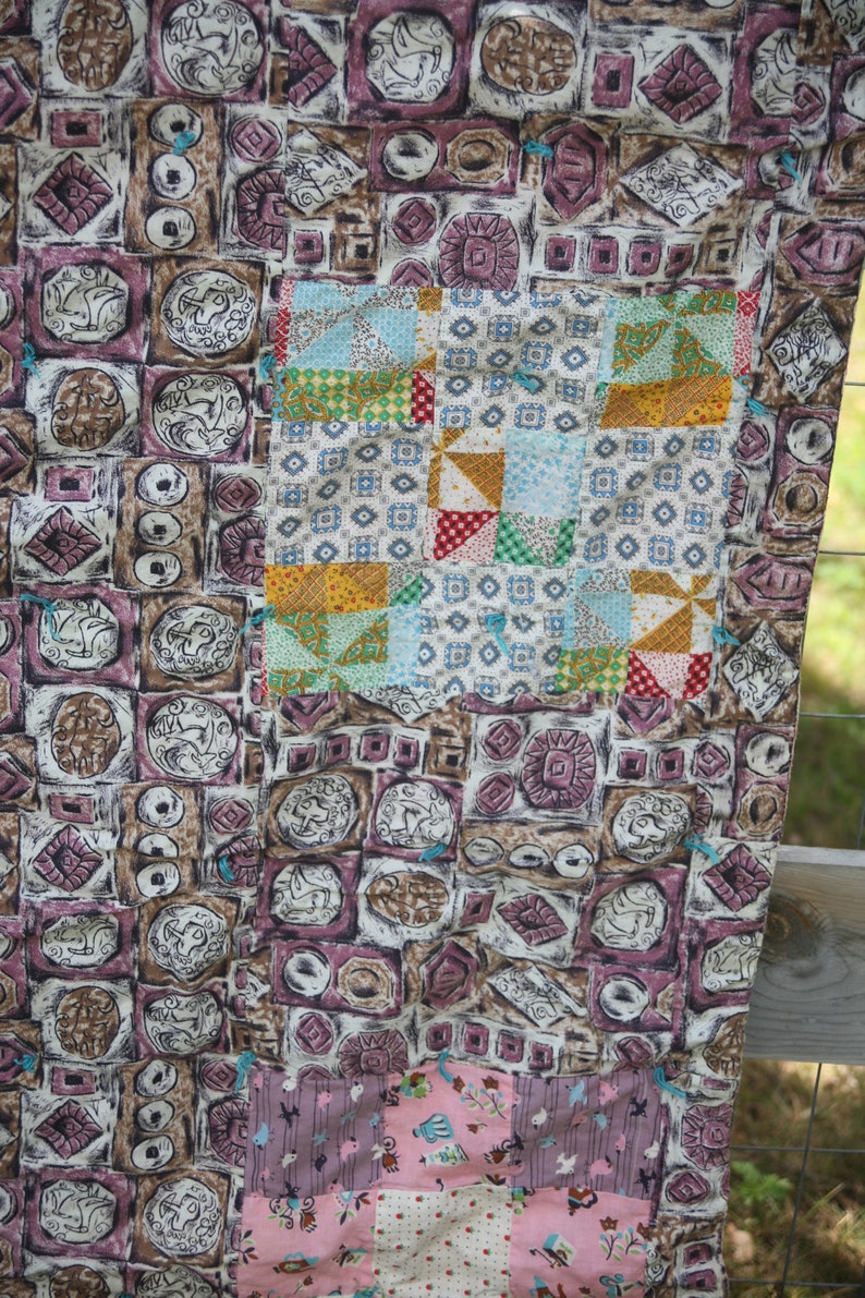 May include: A patchwork quilt with a mix of patterned fabrics. The main section features repeating square designs in brown, purple, and cream. A smaller section has colorful squares and triangles. The quilt is a mix of vintage and modern styles.