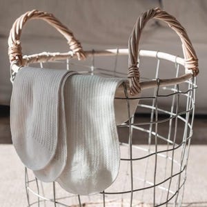 May include: A metal wire basket with wicker handles contains two pairs of cream-coloured socks. The socks have a ribbed texture and are draped over the basket's edge. The basket rests on a light surface.