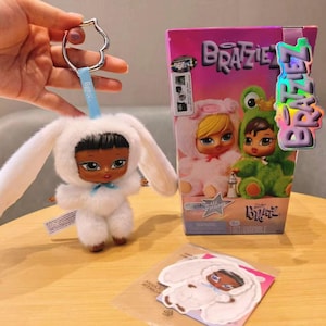 Puede incluir: Una muñeca de peluche blanca con un disfraz de conejito y un lazo azul, sujeta a un llavero. También hay una caja y una pegatina de muñeca Bratz, con muñecas con disfraces de animales. El logotipo de Bratz es visible.