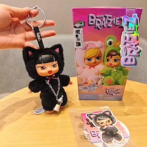 May include: A black Bratz doll keychain in a cat costume with a silver chain and a bottle charm. The doll has a black bob, red lips, and large eyes. A Bratz box and sticker are also visible.