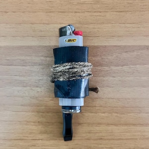 May include: A modified lighter with a black base, wrapped with rope. The lighter is a standard BIC brand, with a white body and the brand logo visible. The top of the lighter is red. The object is on a wooden surface.