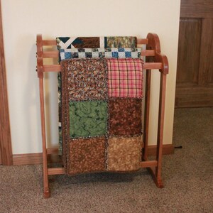 May include: A wooden quilt rack with three quilts hanging on it. The quilts are made of patchwork fabric in various colors, including brown, green, pink, and white.