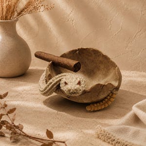 May include: A beige ceramic ashtray shaped like a coiled snake, with a cigar resting inside. The snake has red eyes and a textured body. The ashtray is on a neutral-toned surface, with a vase and dried plants in the background.