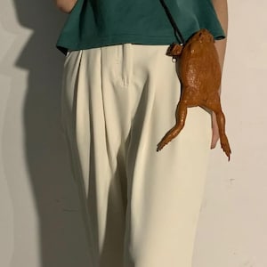 Może przedstawiać: Brązowa skórzana torba na ramię w kształcie żaby z długim paskiem. Torba ma szczegółowe nogi i teksturowaną powierzchnię i jest noszona przez osobę ubraną w zielony top i kremowe spodnie.