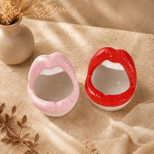 Può includere: Due ciotole in ceramica a forma di bocca aperta, una rifinita con smalto lucido rosa chiaro e l'altra con un vivace smalto rosso lucido. Entrambe le ciotole presentano dettagli di denti bianchi lungo il bordo interno superiore e una base.