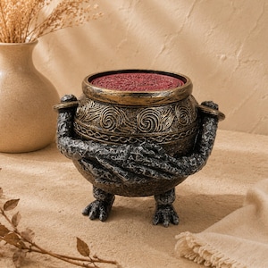 May include: A decorative, fantasy-themed pot with a red interior and intricate gold and bronze detailing. The pot is held by two arms and stands on two feet, creating a unique and artistic design.