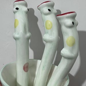 May include: Three whimsical ceramic spoons with a white base, light green accents, and red and black details. Each spoon features a yellow circle and a unique, stylized design. The spoons are displayed in a white bowl.