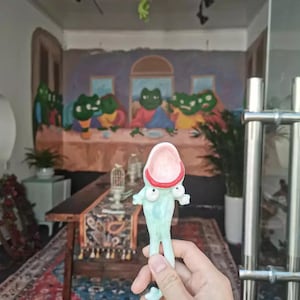 May include: A light blue frog-shaped spoon with a wide open mouth and a red rim. The spoon has large, round eyes and small arms. The background features a mural of frogs at a table, reminiscent of The Last Supper.