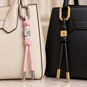 May include: Two bag charms attached to handbags. One charm features a pink cord with a silver-toned metal cube engraved with the letter M, white beads, and silver-toned end caps. The other features a black cord with a gold-toned cube engraved with the letter A and gold-toned end caps.