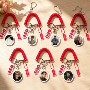 May include: Seven keychains with red coiled cords, each featuring a different image of a person. Each keychain has a small red tag with white text and a charm with three red and white circles. The keychains are on a light brown surface.