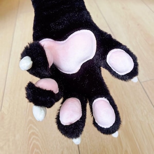 Peut inclure: Une patte noire en peluche avec des coussinets roses et des griffes blanches. La fausse fourrure est douce et duveteuse, avec un design détaillé. La patte fait probablement partie d'un costume ou d'un article fantaisie.