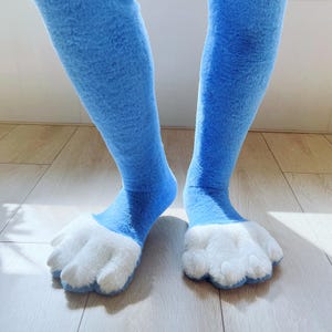 May include: A pair of blue furry leg warmers with white paw-shaped feet. The leg warmers are a solid light blue color and have a soft, plush texture. The feet are white and have a paw-like design.