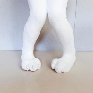 May include: A pair of white plush animal legs featuring soft, textured fabric and padded feet with distinct toes and small, rounded claws. The legs are positioned upright, showcasing the furry material and the detailed construction of the paws.