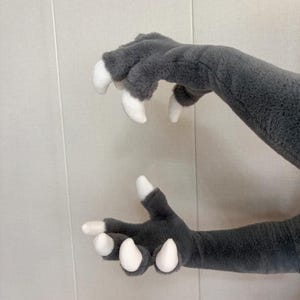 Könnte beinhalten: Ein Paar graue Plüschhandschuhe mit langen Ärmeln und vier Fingern an jeder Hand. Jeder Finger ist mit einer glatten, weißen, gebogenen Kralle versehen. Der Stoff hat eine weiche, flauschige Textur und das Design ahmt das Aussehen von Tierpfoten nach.