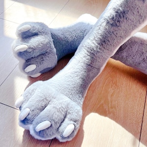 Peut inclure: Une paire de pattes de chat en peluche gris clair avec une longue et douce fourrure et des griffes blanches. Les pattes sont posées sur un sol en bois, l'une chevauchant l'autre. Les pattes en peluche sont un article fantaisiste et ludique.
