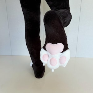 May include: A pair of black plush leggings featuring a white faux fur paw design on the sole of one foot. The paw pad is accented with soft pink, rounded, padded sections that mimic the appearance of an animal foot, complete with white fur.