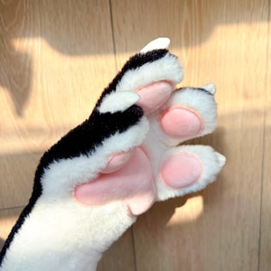 Peut inclure: Gros plan d'une patte en peluche avec des détails noirs, blancs et roses. La patte présente une section supérieure noire et blanche, de la fourrure blanche et des coussinets roses. Les griffes sont blanches.