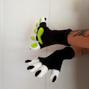 Könnte beinhalten: Ein Paar fingerlose Handschuhe aus schwarzem Kunstfell. Ein Handschuh hat leuchtend hellgrüne Pfotenballen und weiße Fellakzente an den Fingern. Der andere Handschuh hat weiße Fellfingerspitzen mit schwarzen, krallenartigen Details an den Enden.