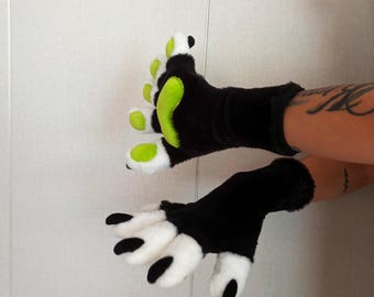 Op de afbeelding: Een paar vingerloze handschoenen van zwart imitatiebont. Eén handschoen heeft fel limoengroene pootkussentjes en witte bontaccenten op de vingers. De andere handschoen heeft witte vingertoppen van bont met zwarte klauwachtige details aan de uiteinden.