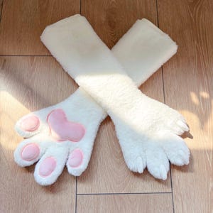 Könnte beinhalten: Ein Paar weiße Plüsch-Armstulpen mit rosa Pfotenpolstern. Die Armstulpen sind flauschig und weich, mit einem herzförmigen Polster in der Mitte der Pfote. Die Pfoten haben fünf rosa Zehenpolster.
