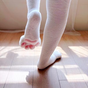 May include: A pair of white plush tights featuring integrated foot covers designed to resemble animal paws. The foot sections are covered in soft white faux fur and include raised, rounded pink pads on the soles to mimic paw prints.