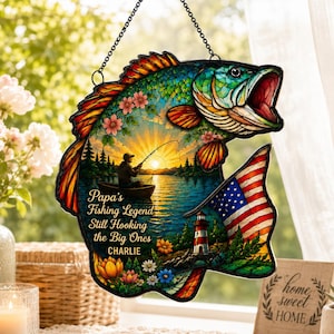 May include: A colorful, fish-shaped hanging decoration with a fishing scene and an American flag design. The text reads "Papa's Fishing Legend Still Hooking the Big Ones CHARLIE". The decoration is suspended by a chain.