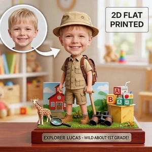 May include: A custom bobblehead figure of a child dressed as an explorer in a tan outfit, hat, and backpack. The figure stands on a wooden base with the text EXPLORER LUCAS - WILD ABOUT 1ST GRADE! It includes a giraffe, magnifying glass, and blocks.