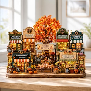 May include: A miniature autumn village display featuring shops named Cookie Shop, Pie Parlor, Harvest Goods, Tea Shop, Candle Stand, and Floral Cottage. A central tree with orange leaves stands behind a banner reading Amber Festival.