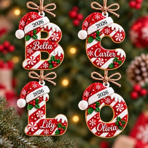 May include: Four hanging Christmas ornaments shaped like the letters B, C, L, and O. Each letter features a red and white candy cane stripe pattern, a Santa hat with the year 2026, green holly leaves, and a personalized name in white script.