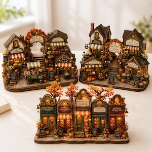 May include: Three miniature illuminated village structures featuring autumn-themed storefronts like Maple Market Square, Harvest Festival, and Hearth & Gold Bakery. Each building displays intricate details, warm interior lighting, and fall foliage.