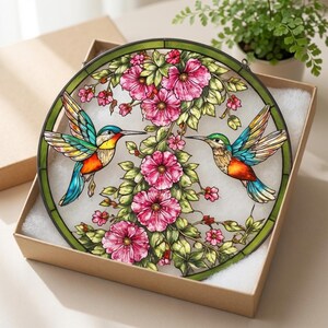 Puede incluir: Una vidriera redonda con dos colibr&iacute;es de alas y picos coloridos, cerca de flores rosas y hojas verdes. La obra de arte est&aacute; enmarcada por un borde verde y se presenta en una caja de cart&oacute;n.