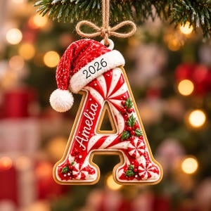 May include: A letter A Christmas ornament shaped like a cookie with red and white peppermint swirl icing, decorated with holly berries and leaves. It features the name Amelia and the year 2026, topped with a miniature red Santa hat and twine.