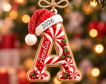 May include: A letter A Christmas ornament shaped like a cookie with red and white peppermint swirl icing, decorated with holly berries and leaves. It features the name Amelia and the year 2026, topped with a miniature red Santa hat and twine.
