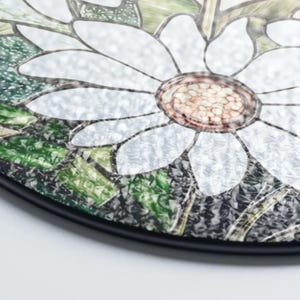 Puede incluir: Decoraci&oacute;n redonda de vidriera con dise&ntilde;o de margarita. La flor tiene p&eacute;talos blancos y centro marr&oacute;n, con hojas verdes y borde negro. El vidrio tiene un acabado texturizado. Adecuado para la decoraci&oacute;n del hogar.