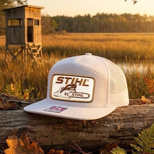 May include: White trucker hat with a mesh back and a rectangular patch featuring the "STIHL" logo and a chainsaw graphic. The hat is resting on a log with fall foliage in an outdoor setting.