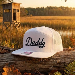 May include: A white trucker hat with the word "Daddy" in black script. The hat is resting on a log with a backdrop of a wooden structure, marshland, and a sunset.