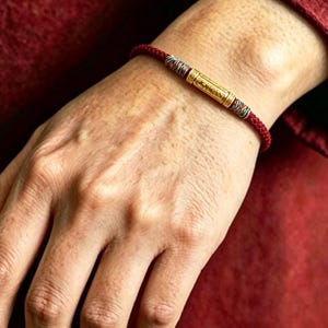 May include: A red braided cord bracelet featuring a central gold-toned cylindrical bead with intricate embossed patterns. The gold bead is flanked by small, multi-colored woven accents on either side, securing the cord in a continuous loop design.