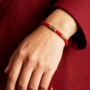 May include: A red braided bracelet featuring gold-coloured beads and accents. The bracelet is worn on a wrist, with the red cord and gold beads contrasting against the skin tone. The bracelet is a simple yet elegant piece of jewellery.
