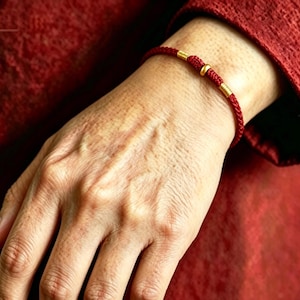 May include: A red braided cord bracelet featuring three small gold-toned cylindrical and spherical metal beads centered on the band. The bracelet is constructed from a thin, woven red string material with a smooth, uniform texture throughout.