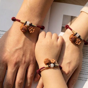 Puede incluir: Tres pulseras a juego con un cordón rojo oscuro, una cuenta de madera marrón tallada, un pequeño dije de flor de madera de cinco pétalos y una cuenta blanca translúcida. Cada pulsera está asegurada con nudos ajustables y detalles de hilo.