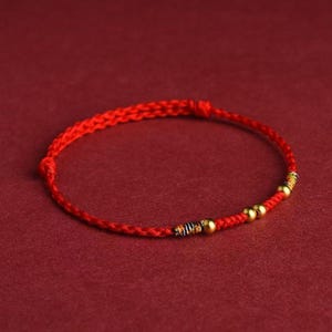 Puede incluir: Una pulsera trenzada roja con cuentas doradas y detalles decorativos. La pulsera está hecha de un material fino y tejido y se muestra sobre un fondo rojo. La pulsera es una pieza de joyería sencilla y elegante.