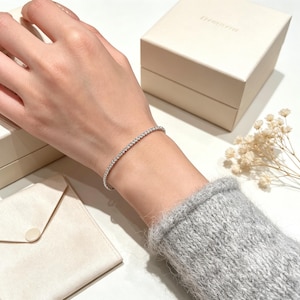 Puede incluir: Una delicada pulsera de tenis en tono plateado que presenta una hilera continua de pequeñas piedras claras y brillantes. La pulsera se lleva en la muñeca, mostrando un diseño de banda metálica fina y flexible que rodea el brazo con elegancia.