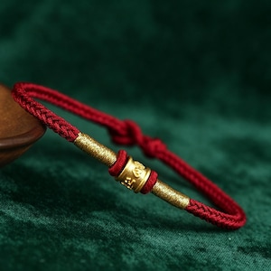 May include: A red braided bracelet with gold-coloured cylindrical accents. The bracelet has a knot closure and a central gold bead with etched details. The background is a dark green, contrasting with the bracelet's colours.