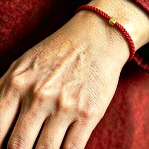May include: A thin red braided cord bracelet featuring a single small, polished gold-colored cube bead centered on the band. The bracelet is constructed from a smooth, woven material and sits securely around the wrist, showcasing a minimalist design.