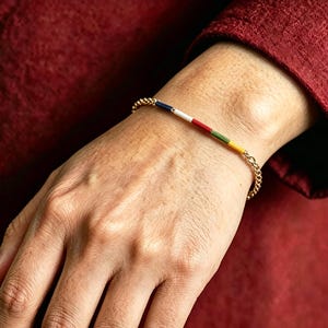 May include: A gold chain bracelet with a series of colourful beads. The beads are arranged in a linear pattern, featuring white, red, green, and yellow. The bracelet is worn on a wrist, with a red fabric background.