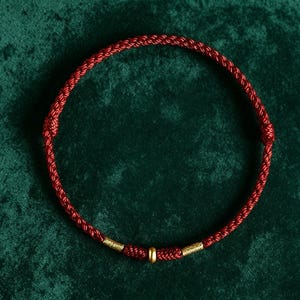 May include: A circular bracelet made of deep red braided cord. The design features two small knots on the sides and a central section adorned with a smooth gold-toned bead flanked by two textured gold-toned cylindrical accents along the cord.