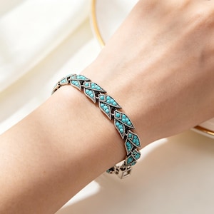 May include: A silver-toned metal bracelet featuring a series of connected leaf-shaped links. Each leaf link is inlaid with small, bright turquoise-colored stones arranged in a textured pattern. The bracelet has a secure clasp closure.