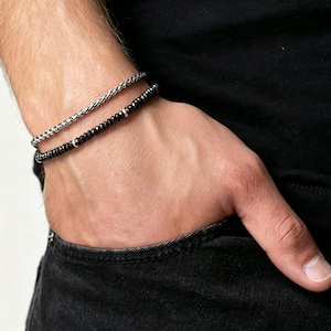 Può includere: Un set di due bracciali indossati insieme. Un bracciale presenta un design a catena intrecciata color argento. Il secondo bracciale è composto da piccole perline nere rotonde, intervallate da distanziatori in metallo color argento.