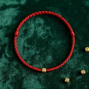 May include: A red braided cord bracelet featuring a single small gold-toned cube bead centered on the band. The bracelet is circular in shape and is accompanied by three additional loose gold-toned cube beads placed nearby on the surface.