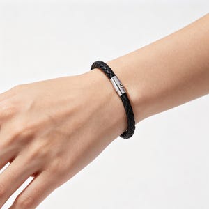May include: A black braided leather bracelet featuring a cylindrical silver-toned metal magnetic clasp. The bracelet consists of multiple thin leather strands woven together into a single cord, secured by a polished metallic locking mechanism.